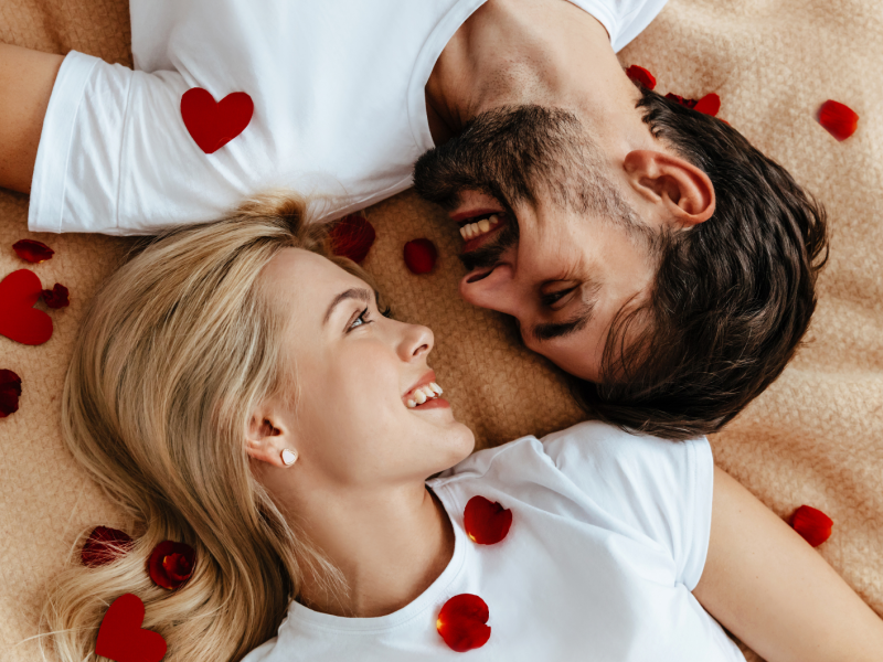 Ein Mann und eine Frau, die auf einer Decke gemeinsam liegen und sich zwischen Rosenblättern verliebt anschauen.