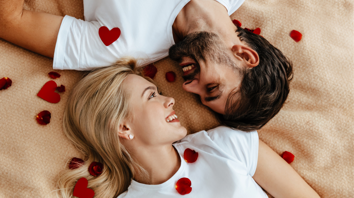 Ein Mann und eine Frau, die auf einer Decke gemeinsam liegen und sich zwischen Rosenblättern verliebt anschauen.