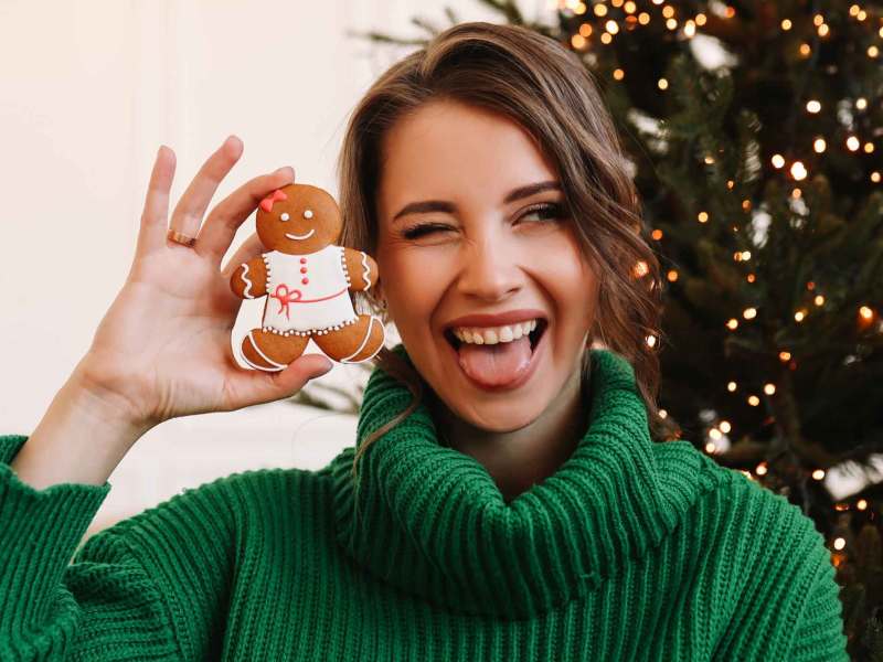 Frau mit grünem Pullover hält einen Lebkuchenmann in der Hand und zwinkert dabei. Im Hintergrund steht ein Weihnachtsbaum.