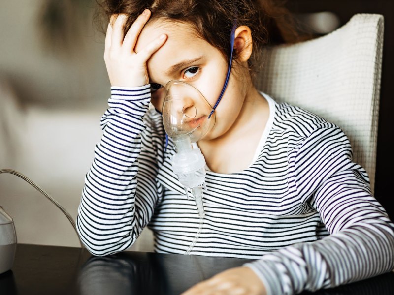 Mädchen, ca. 6 Jahre, sitzt am Tisch und trägt eine Inhalationsmaske, daneben steht das Inhalationsgerät.