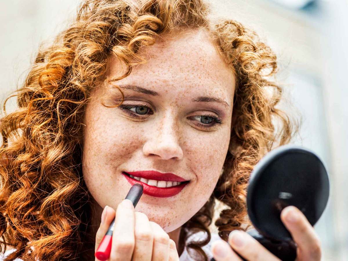 Frau mit roten Locken schminkt sich die Lippen mit einem roten Lippenstift.