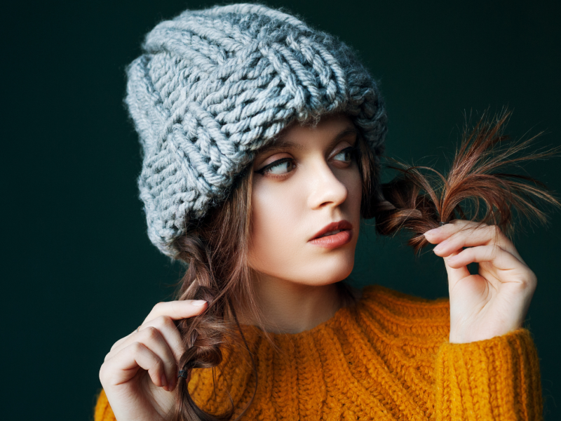 Eine junge Frau in einem gelben Strickpullover und einer grob gestrickten grauen Mütze. Sie hält spielerisch ihre braunen Haarspitzen mit den Händen, während sie seitlich schaut. Der Hintergrund ist dunkelgrün.