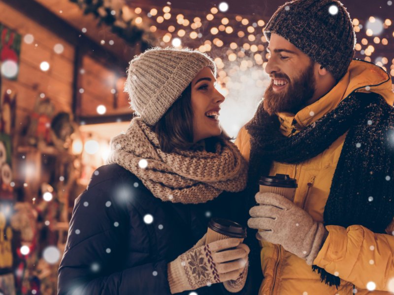 Frau und Mann in Winterkleidung auf dem Weihnachtsmarkt lächeln sich verliebt an