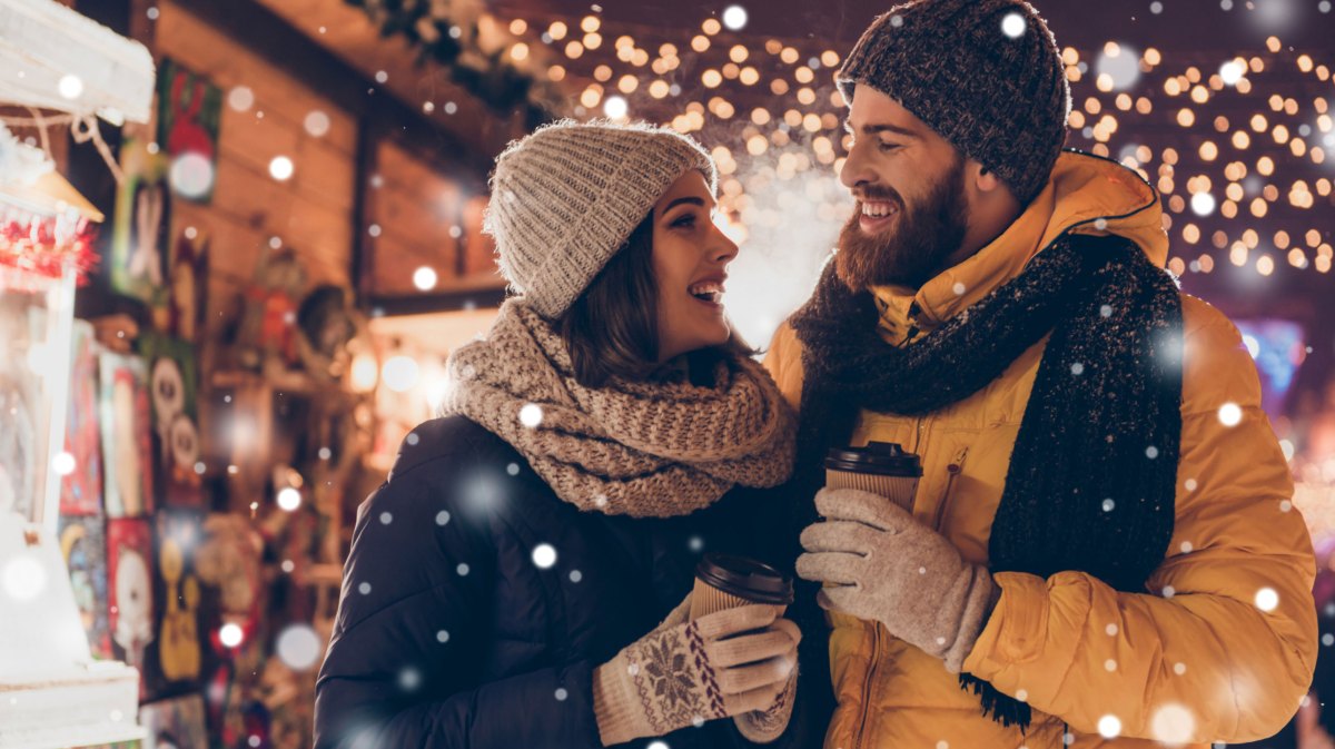 Frau und Mann in Winterkleidung auf dem Weihnachtsmarkt lächeln sich verliebt an