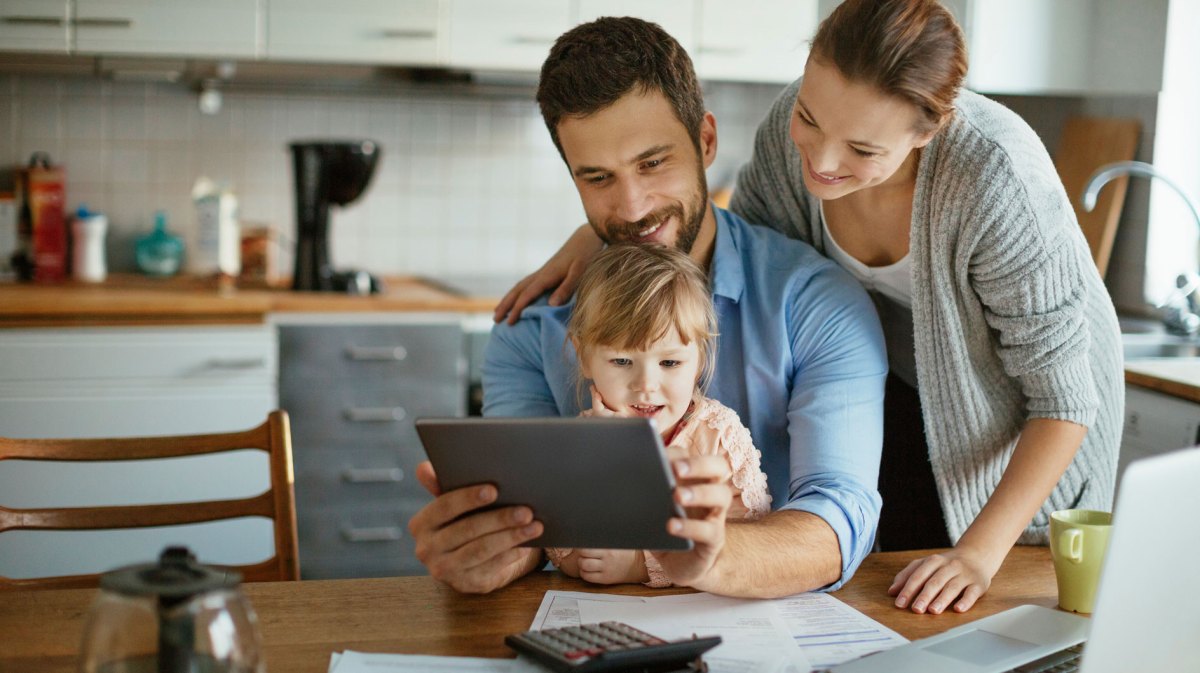 Dreiköpfige Familie am Küchentisch schaut sich wohlwollend etwas auf einem Tablet an. Um sie herum liegen Rechnungen und ein Taschenrechner.