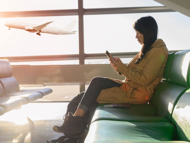 Frau am Handy sitzt am Flughafen, Flugzeug startet im Hintergrund