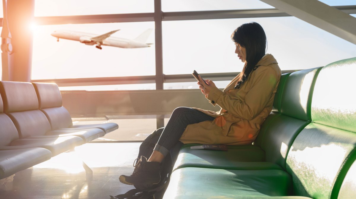 Frau am Handy sitzt am Flughafen, Flugzeug startet im Hintergrund