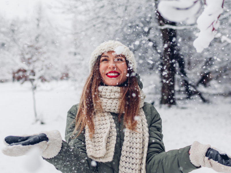 Junge Frau mit roten Lippen, Wollmütze und Schal lacht strahlend in fallendem Schnee vor winterlicher Parklandschaft.