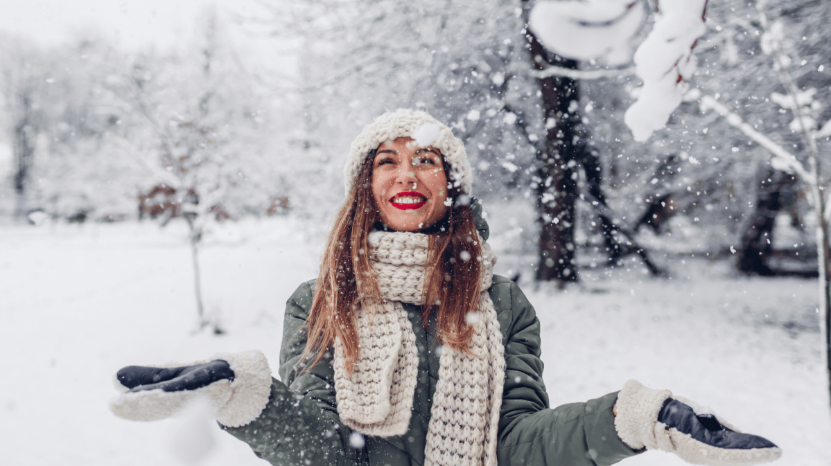 Junge Frau mit roten Lippen, Wollmütze und Schal lacht strahlend in fallendem Schnee vor winterlicher Parklandschaft.