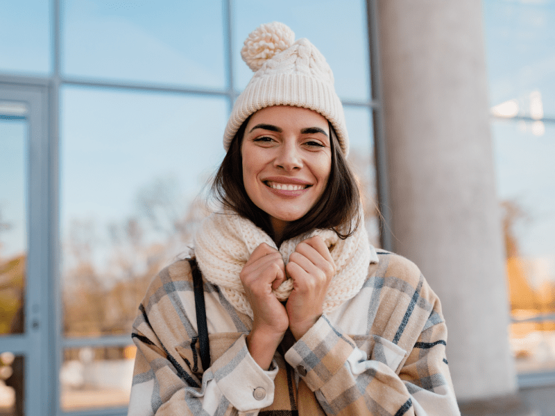 Fröhliche Frau mit beigem Winteroutfit hält Schal fest, sonniger Tag, reflektierende Fenster im Hintergrund.