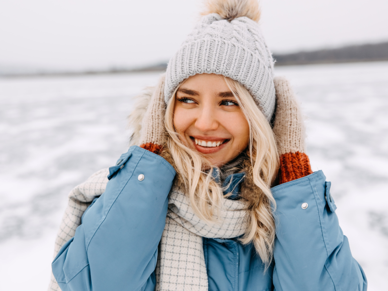 Frau im Schnee, mit Winterjacke, Mütze und Schal, die sich an die Ohren packt und zur Seite lächelt.