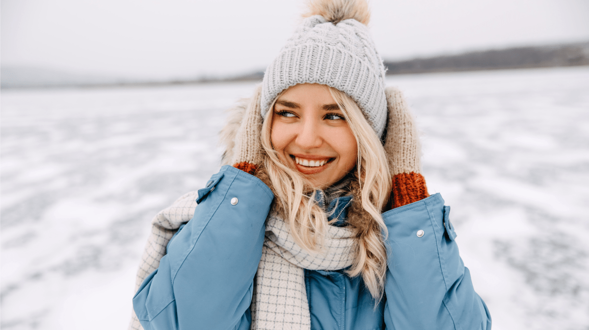 Frau im Schnee, mit Winterjacke, Mütze und Schal, die sich an die Ohren packt und zur Seite lächelt.