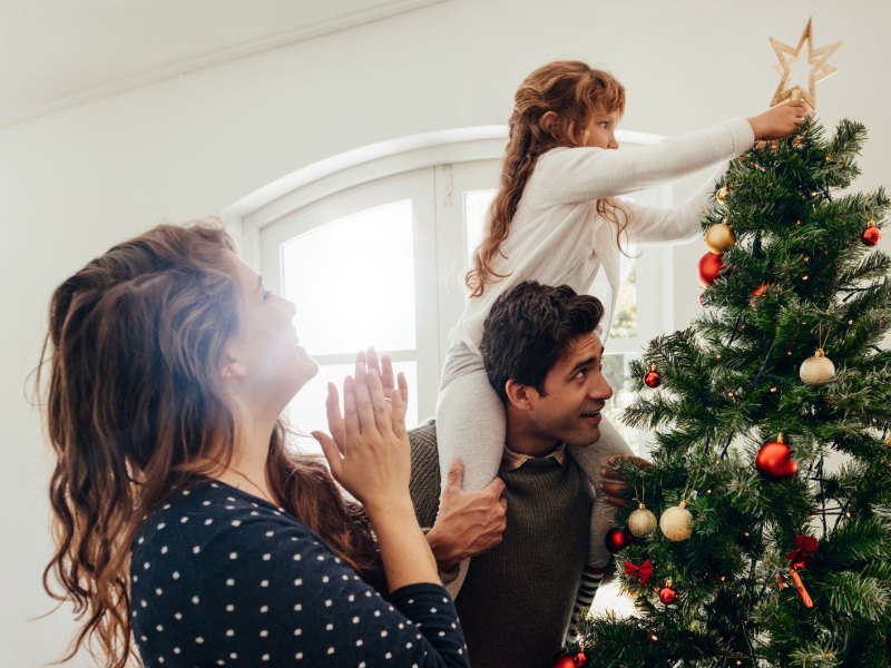 Mutter, Vater und Tochter haben den Weihnachtsbaum geschmückt. Die Tochter, auf den Schultern des Vaters sitzend, steckt dem Baum die Stern-Spitze an.