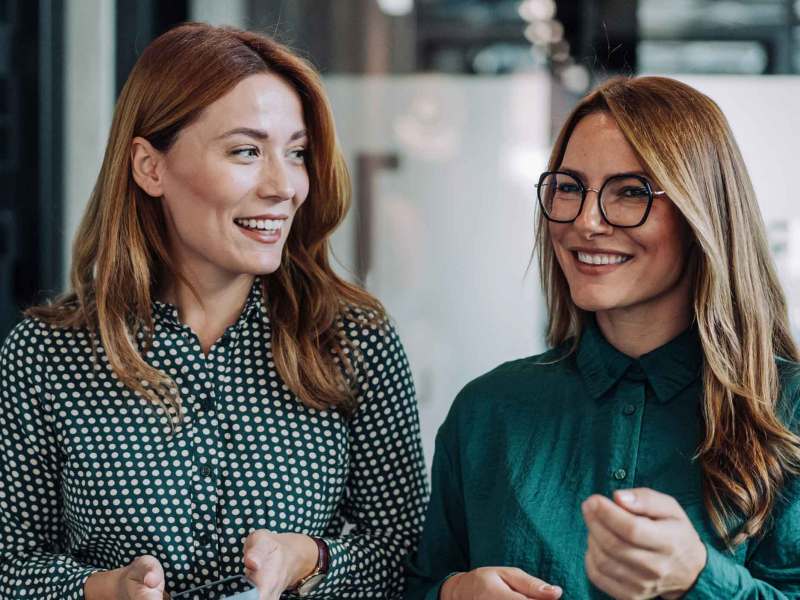 Zwei erfolgreiche junge Frauen mit Businesskleidung gehen durch das Büro und unterhalten sich angeregt.