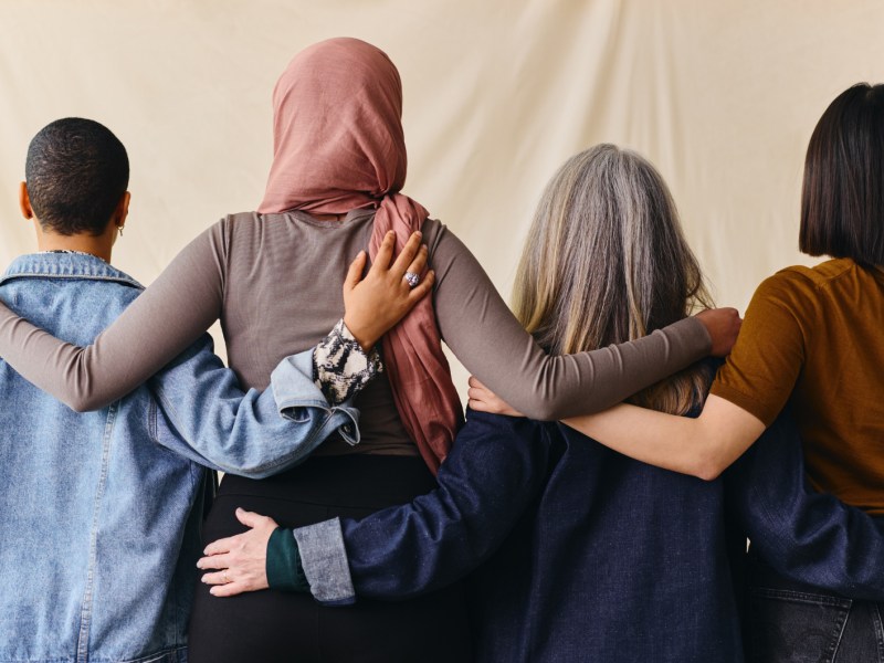 Vier Frauen stehen mit Rücken zur Kamera, haben ihre Arme um die Schultern gelegt.