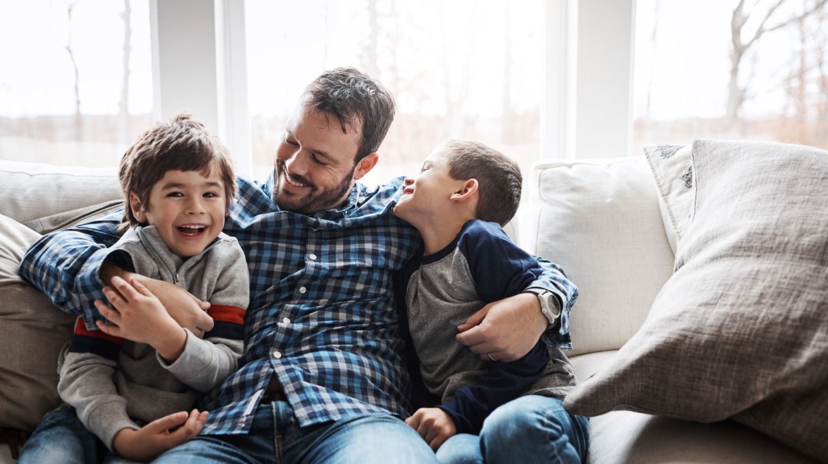 Vater sitzt auch der Couch, seine Söhne rechts und links von ihm. Er hat seine Arme um die Jungs gelegt und alle lachen.