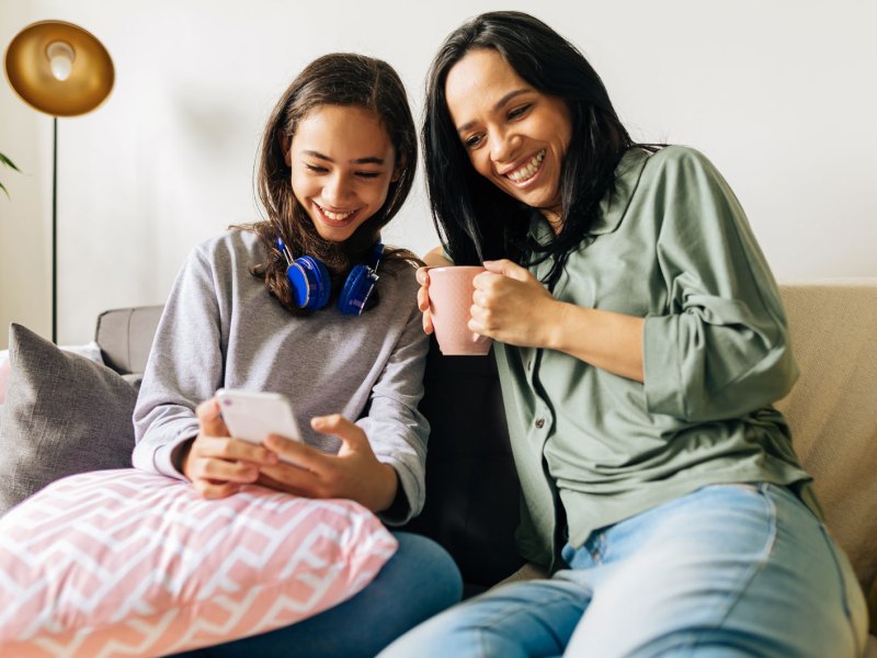 Mutter und Tochter sitzen auf dem Sofa. Die Tochter zeigt der Mutter etwas auf ihrem Smartphone. Beide wirken sehr vertraut und entspannt miteinander.