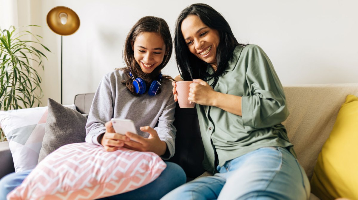 Mutter und Tochter sitzen auf dem Sofa. Die Tochter zeigt der Mutter etwas auf ihrem Smartphone. Beide wirken sehr vertraut und entspannt miteinander.