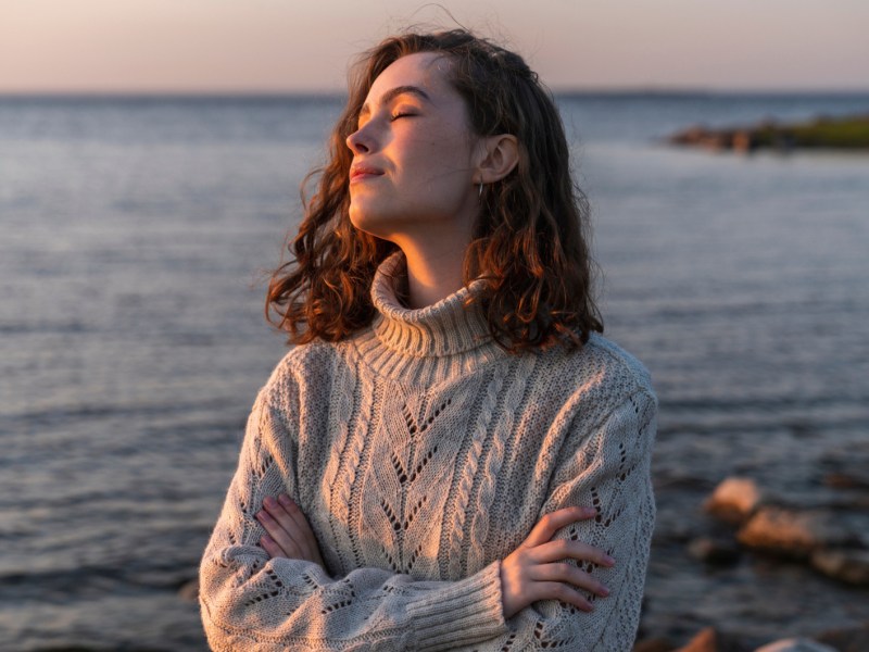 Frau in Strickpullover steht an Küste und hat Gesicht zum Sonnenuntergang gewandt.