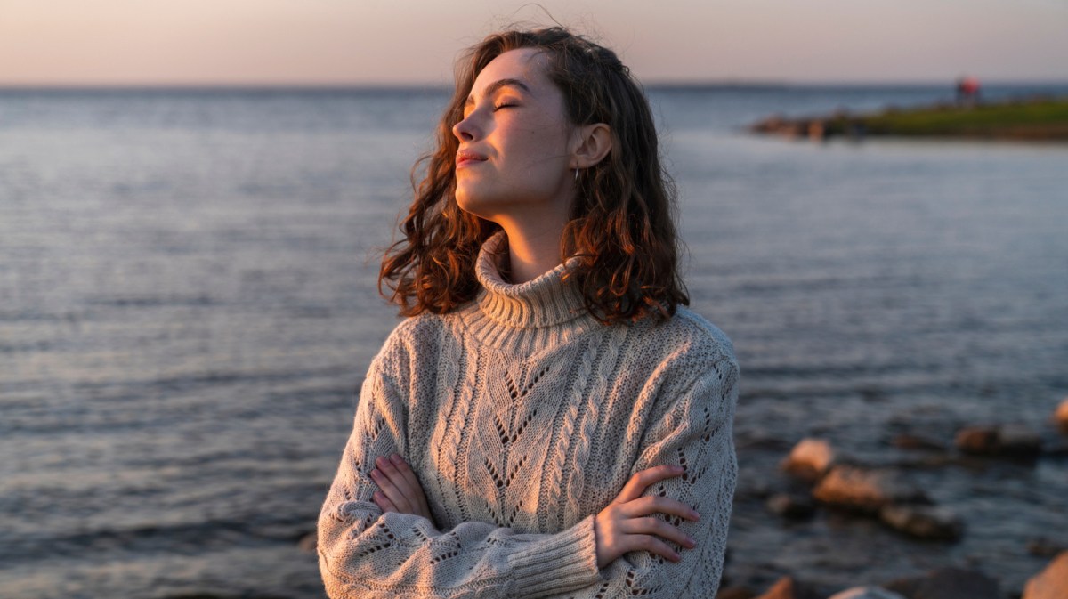 Frau in Strickpullover steht an Küste und hat Gesicht zum Sonnenuntergang gewandt.