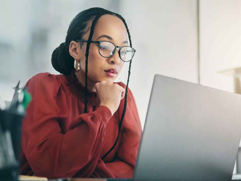 Junge Frau mit dunkelroter Bluse und Brille sitzt nachdenklich vor dem Laptop.