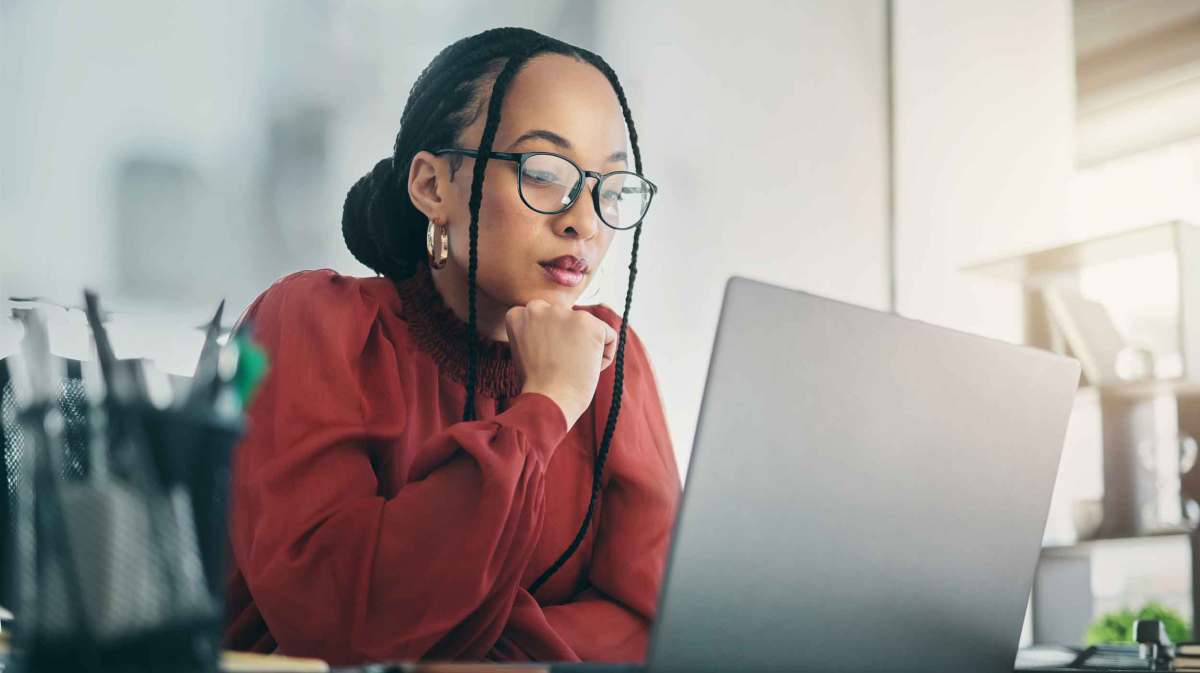 Junge Frau mit dunkelroter Bluse und Brille sitzt nachdenklich vor dem Laptop.