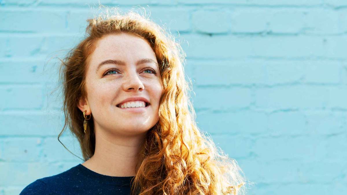 Frau mit roten, lockigen Haaren steht lächelnd vor einer hellblauen Wand.