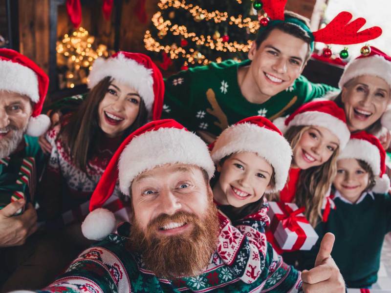 Selfie einer Familie, bestehend aus 8 Personen, die alle Weihnachtspulli und -mütze tragen und vor einem Weihnachtsbaum posieren.