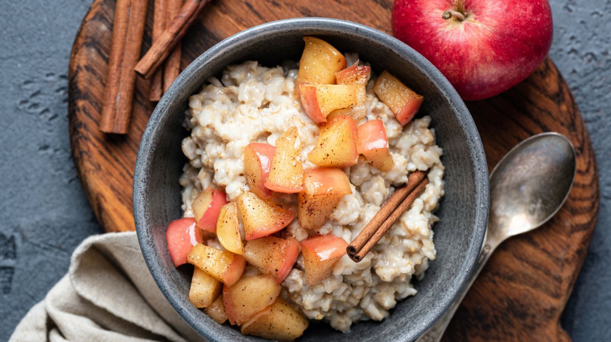 Apfel-Zimt-Porridge in einer schwarzen Schüssel auf einem Holzbrett. Ein Apfel, ein Löffel & Zimtstangen liegen daneben.