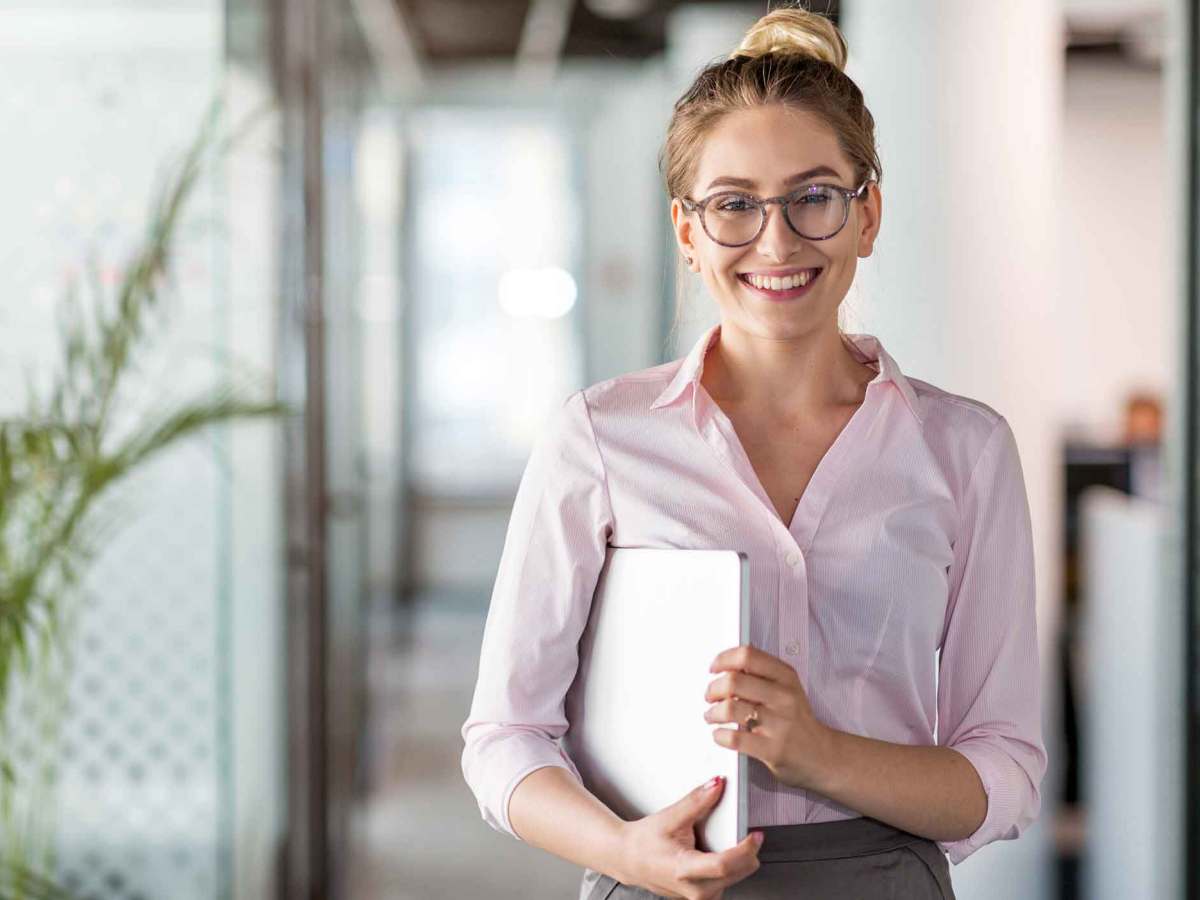 Frau mit Brille steht im Büro und hält ein Notebook unter dem Arm.