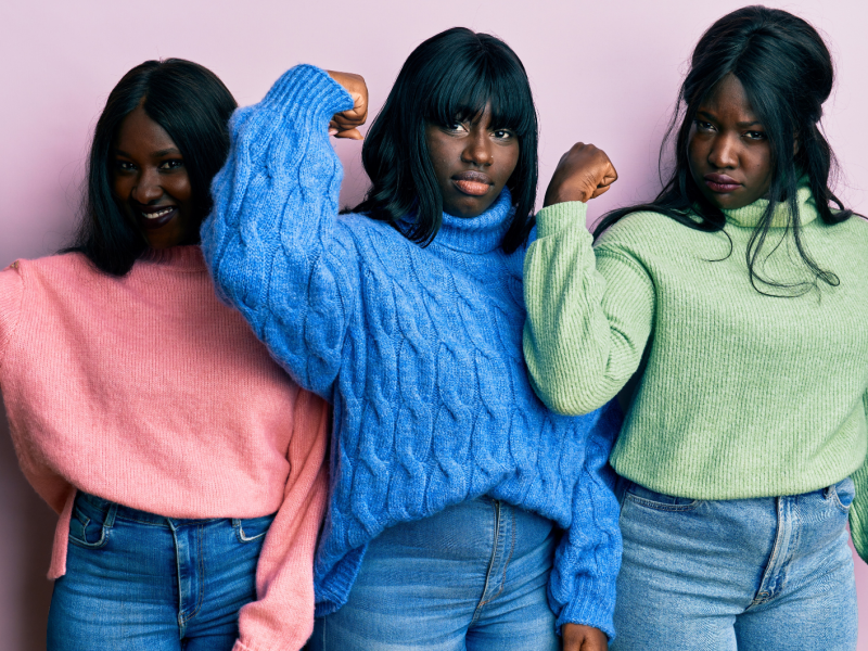 Drei junge Frauen mit schwarzem Haar posieren entschlossen vor rosa Hintergrund, tragen Strickpullover in Pink, Blau und Grün.