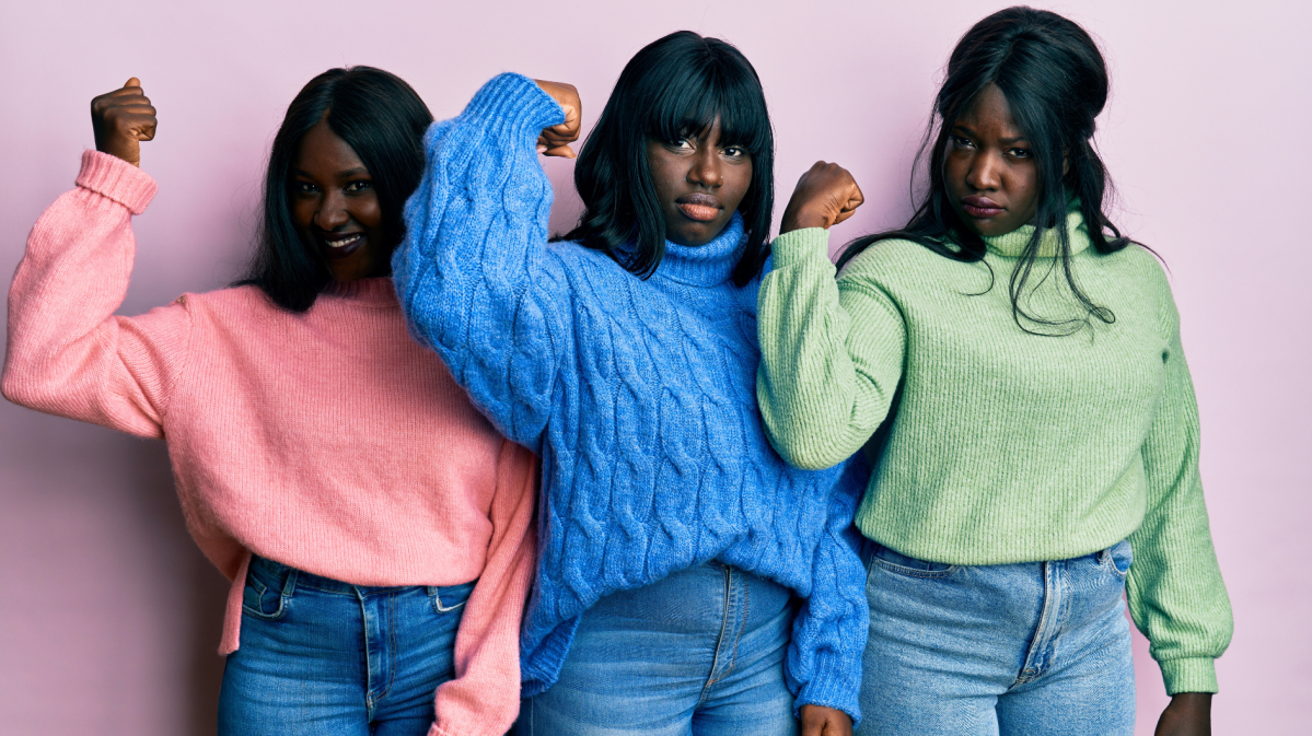 Drei junge Frauen mit schwarzem Haar posieren entschlossen vor rosa Hintergrund, tragen Strickpullover in Pink, Blau und Grün.