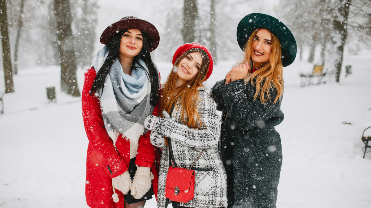 Drei Frauen im Schnee, mit Wintermantel und Hüten, die allesamt in die Kamera lächeln, während es um sie herum schneit.