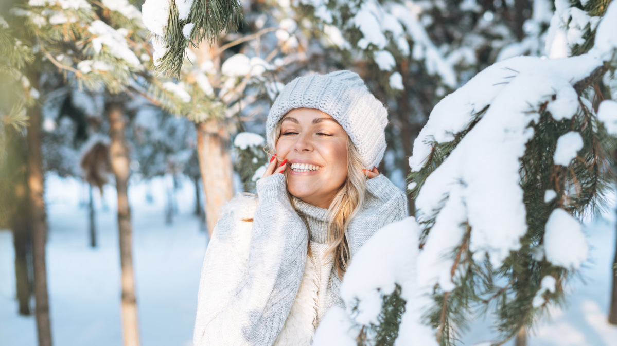 Junge Frau mit strahlendem Lächeln und rotem Nagellack lehnt sich im warmen Winter-Outfit an einen schneebedeckten Baum.