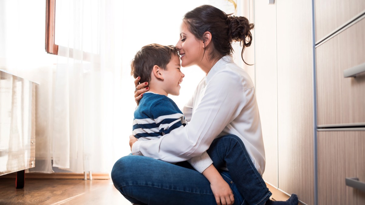 Sohn, ca. 6 Jahre alt, sitzt auf dem Fußboden auf dem Schoß seiner Mutter und sie gibt ihm einen Kuss auf die Stirn.