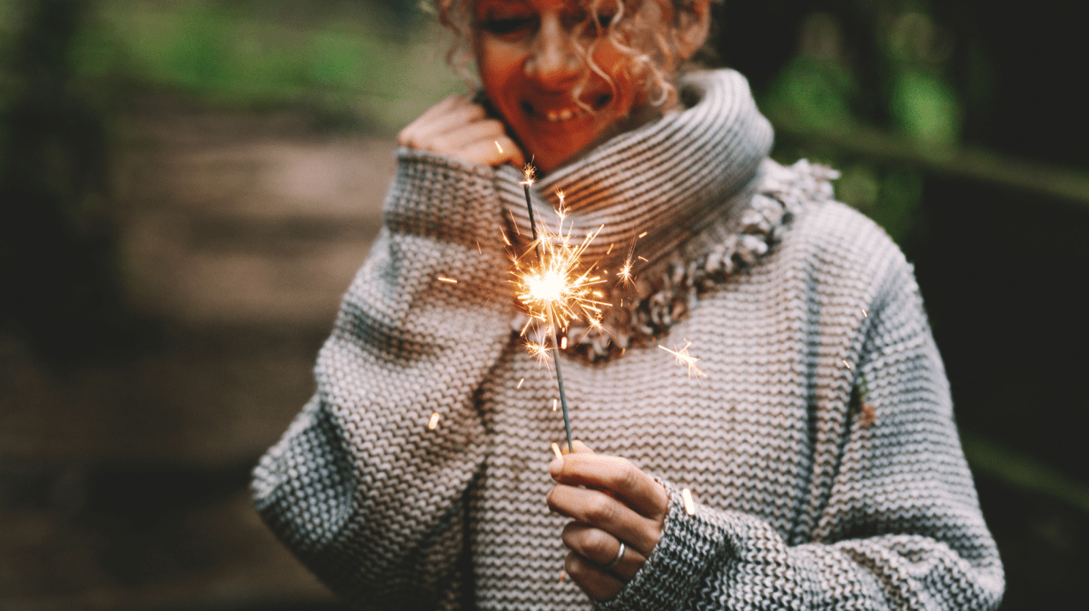 Eine lachende junge Frau mit lockigem blondem Haar hält eine brennende Wunderkerze, trägt einen groben Wollpullover im Wald.