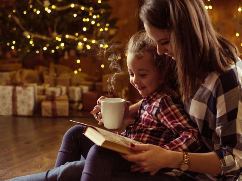 Mutter und Tochter sitzen gemütlich im Wohnzimmer und lesen ein Buch zusammen. Hinter ihnen leuchtet der Weihnachtsbaum.