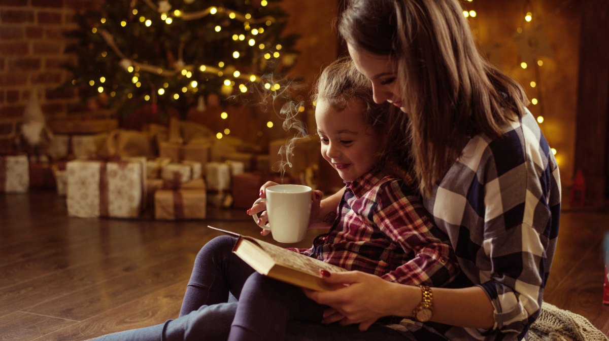 Mutter und Tochter sitzen gemütlich im Wohnzimmer und lesen ein Buch zusammen. Hinter ihnen leuchtet der Weihnachtsbaum.