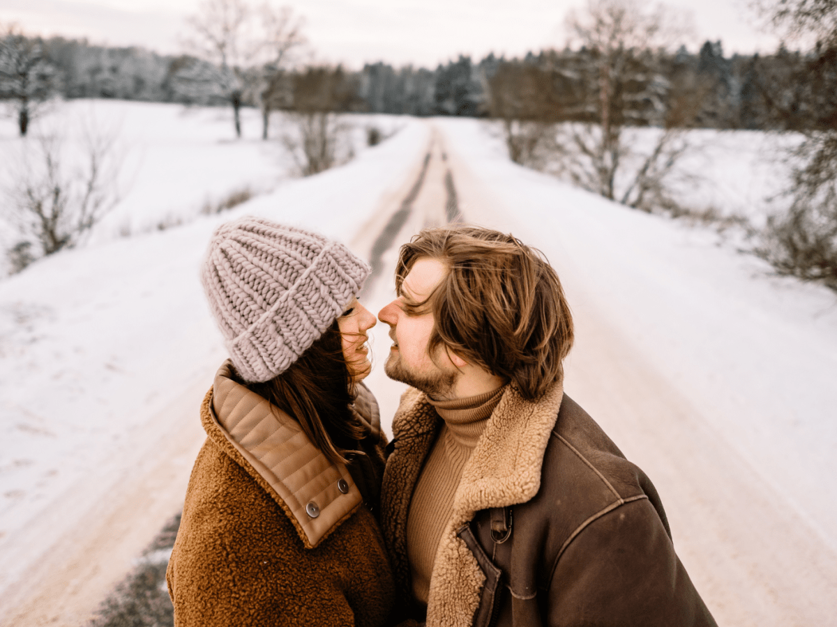 Eine Frau mit Strickmütze und ein Mann in brauner Jacke nähern sich lächelnd in einer schneebedeckten Landstraße an.