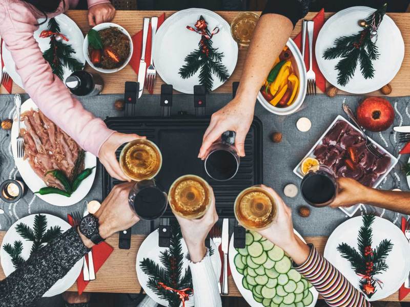 Familie sitzt am Holztisch mit Raclette-Grill in der Mitte. Alle stoßen an.