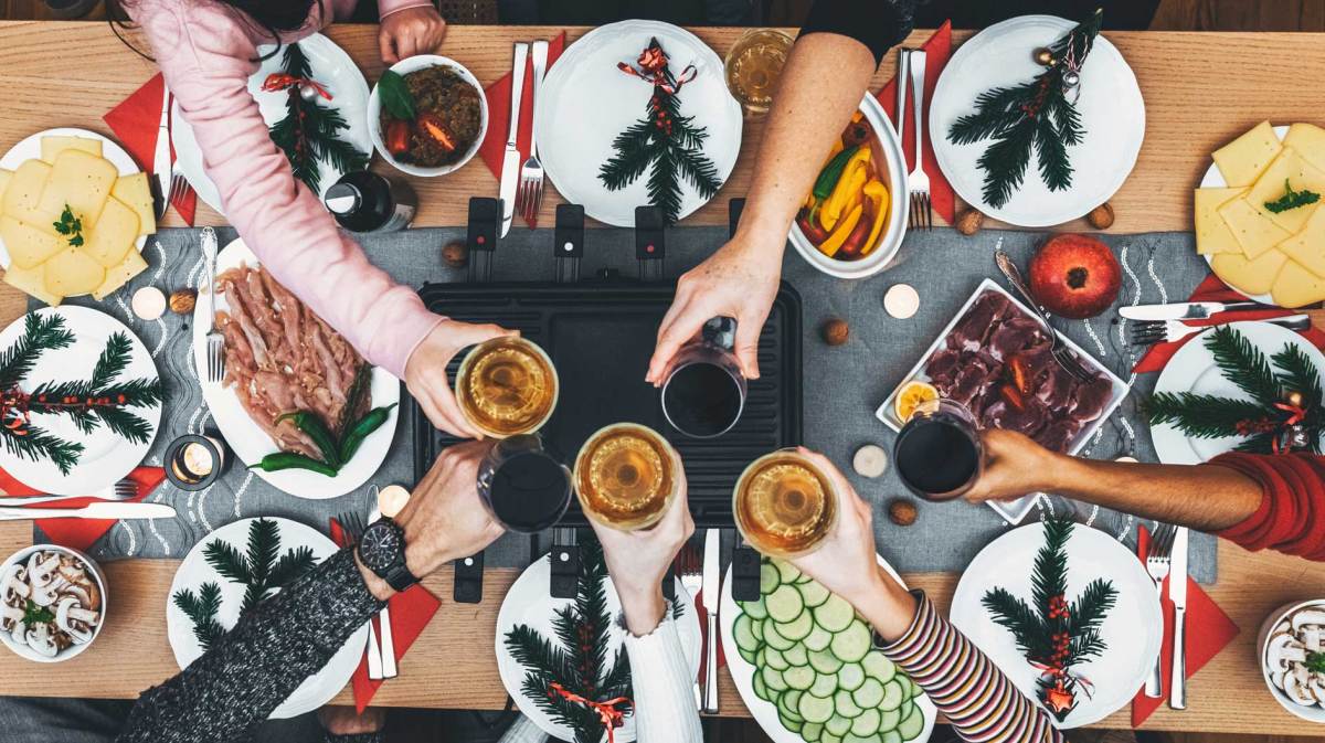 Familie sitzt am Holztisch mit Raclette-Grill in der Mitte. Alle stoßen an.