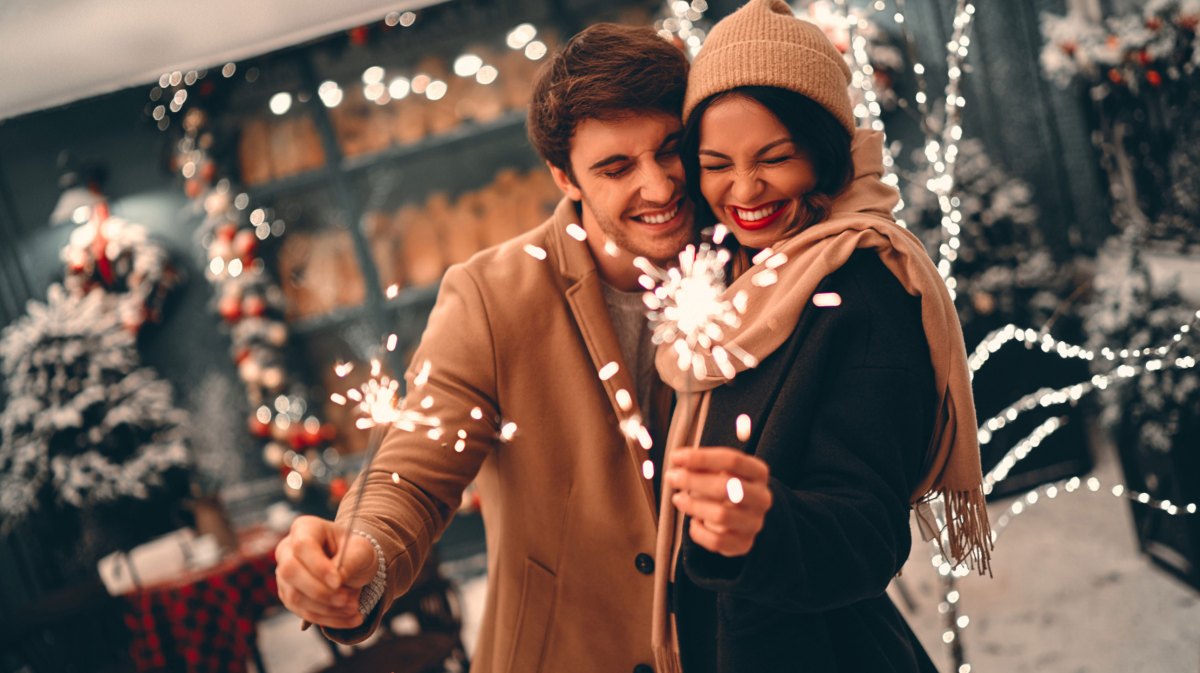 Mann und Frau in Winterbekleidung vor weihnachtlich geschmücktem Haus, mit Wunderkerzen in der Hand.