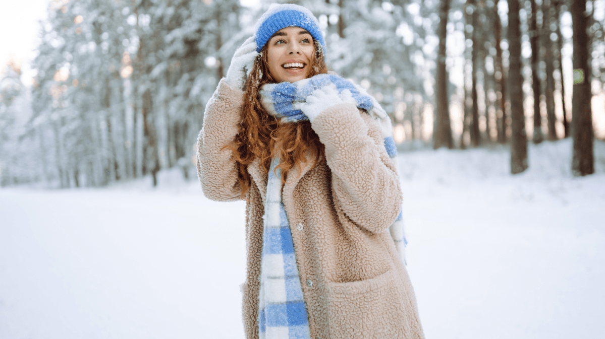 Junge Frau mit braunen Locken und blauer Mütze lacht fröhlich im verschneiten Winterwald, eingehüllt in einen beigen Mantel und karierten Schal.