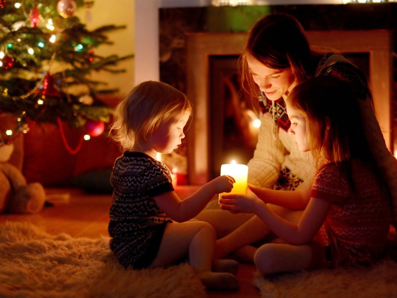 Mutter und Töchter sitzen auf dem Fußboden neben ihrem Weihnachtsbaum und halten gemeinsam eine einzige Kerze.