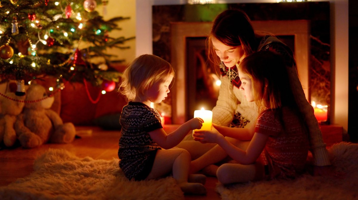 Mutter und Töchter sitzen auf dem Fußboden neben ihrem Weihnachtsbaum und halten gemeinsam eine einzige Kerze.