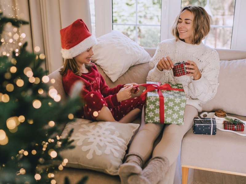 Mutter und Tochter sitzen in gemütlichen Sachen auf dem Sofa, neben ihnen leuchtet der Weihnachtsbaum und sie packen Geschenke aus.