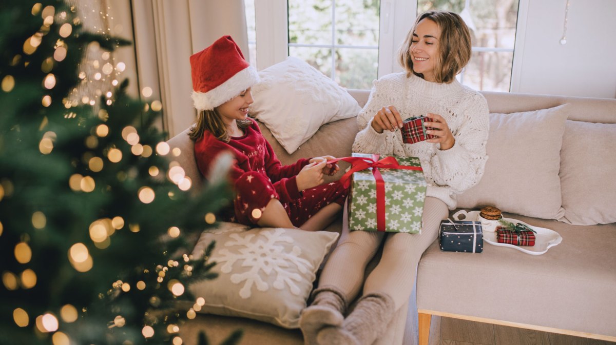 Mutter und Tochter sitzen in gemütlichen Sachen auf dem Sofa, neben ihnen leuchtet der Weihnachtsbaum und sie packen Geschenke aus.