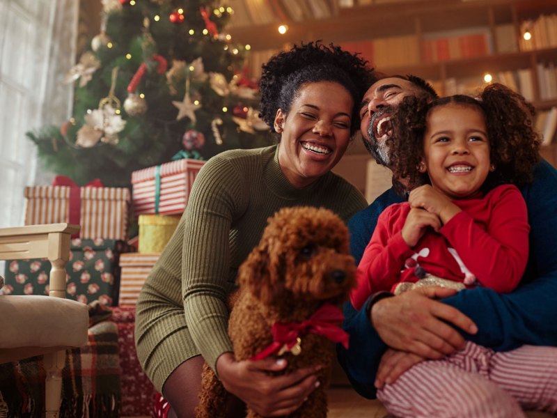 Dreiköpfige Familie posiert mit ihrem Hund vorm Weihnachtsbaum.