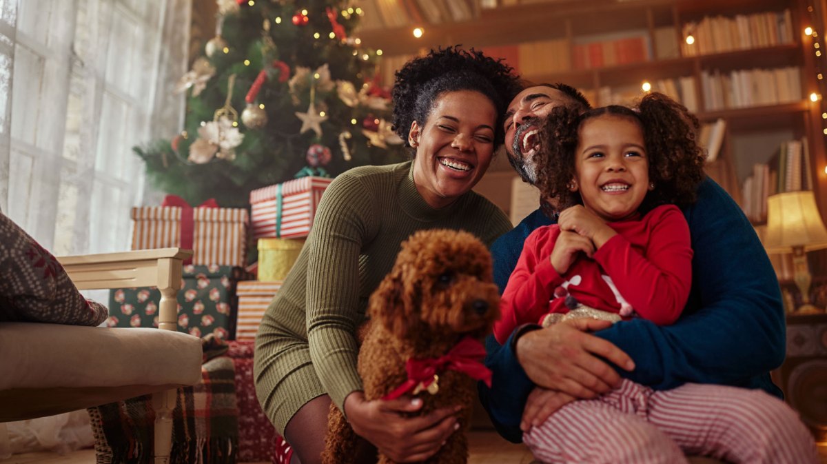 Dreiköpfige Familie posiert mit ihrem Hund vorm Weihnachtsbaum.