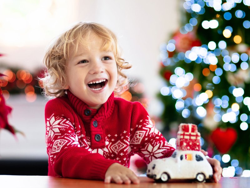 Junge, ca. 6 Jahre, lacht sehr glücklich, hinter ihm steht ein leuchtender Weihnachtsbaum.